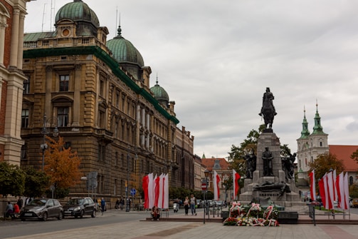 An urban scene featuring a prominent historical monument in a square, surrounded by architectural buildings with detailed facades and domed roofs. Several flags in red and white are displayed around the monument, hinting at a national significance. People are walking and cars are parked along the road, suggesting an active city environment.