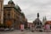 An urban scene featuring a prominent historical monument in a square, surrounded by architectural buildings with detailed facades and domed roofs. Several flags in red and white are displayed around the monument, hinting at a national significance. People are walking and cars are parked along the road, suggesting an active city environment.