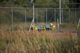 Two benches, one yellow and one green, are positioned alongside a set of yellow stackable chairs on a tennis court. The court is surrounded by a metal chain-link fence and bordered by tall grass and trees in the background, creating a natural and somewhat abandoned appearance.
