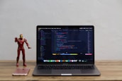 A laptop displaying code on its screen sits on a wooden table. To the left of the laptop, a red and gold superhero action figure stands next to a pink smartphone.