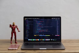 A laptop displaying code on its screen sits on a wooden table. To the left of the laptop, a red and gold superhero action figure stands next to a pink smartphone.