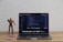A laptop displaying code on its screen sits on a wooden table. To the left of the laptop, a red and gold superhero action figure stands next to a pink smartphone.