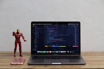 A laptop displaying code on its screen sits on a wooden table. To the left of the laptop, a red and gold superhero action figure stands next to a pink smartphone.
