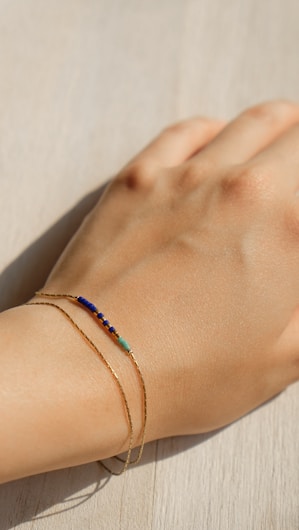 A delicate gold bracelet is worn on a wrist, featuring small blue and turquoise beads. The hand rests on a light wood surface with natural lighting casting soft shadows.