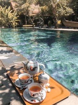 A quaint poolside setting features a tray with two cups of tea, a teapot, a sugar bowl, and a small white flower on a wooden surface next to clear blue water. The background includes lush greenery with palm trees and a wooden structure with a tiled roof.