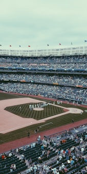A large baseball stadium filled with spectators, featuring a well-maintained baseball field at the center. Players wearing uniforms are gathered near the pitcher's mound, while fans fill the stands, some walking around. The stadium's architecture includes multiple tiers with a row of flags at the top, indicating a lively atmosphere.