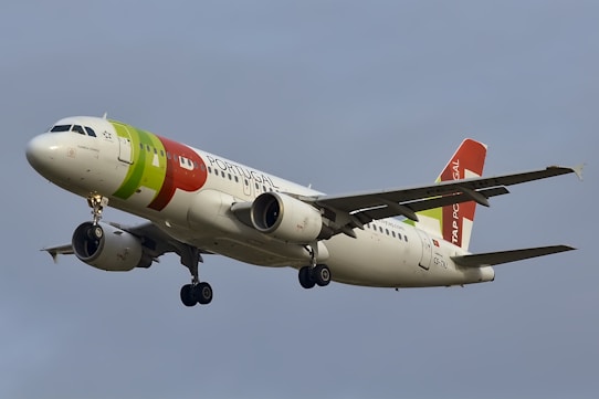 A commercial airplane is flying in the sky with its landing gear extended. The aircraft is painted in white with red, green, and orange accents and the words 'Portugal' and 'TAP' visible on its body and tail.