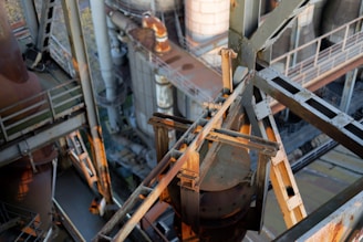 An industrial scene featuring a complex arrangement of metal beams and rusted machinery. The structure includes staircases, pipes, and girders, with sunlight casting shadows and highlighting parts of the equipment.