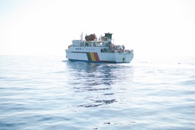 A ferry with a white body and colorful stripes sails on a calm, expansive ocean with clear skies. The vessel has multiple decks and appears to be carrying numerous passengers. The water is a light blue, reflecting the bright sunlight that creates a serene and peaceful atmosphere.