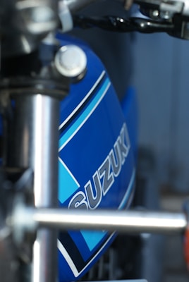 Close-up of a motorcycle with a blue tank featuring the Suzuki logo. The focus is on the tank and handlebars, highlighting the sleek metallic finish and design details. The background is blurred, drawing attention to the motorcycle's branding and construction.