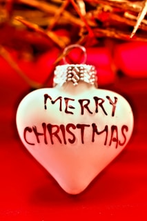 A heart-shaped ornament features the words 'Merry Christmas' in bold letters. The background consists of vibrant red and gold tones with a warm and festive atmosphere, with hints of Christmas decorations.