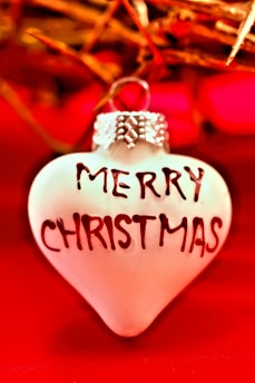 A heart-shaped ornament features the words 'Merry Christmas' in bold letters. The background consists of vibrant red and gold tones with a warm and festive atmosphere, with hints of Christmas decorations.