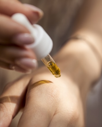A close-up image of a dropper releasing a golden liquid onto the back of a person's hand. The person's skin appears smooth and is being delicately treated with the liquid, conveying a sense of care and attention.