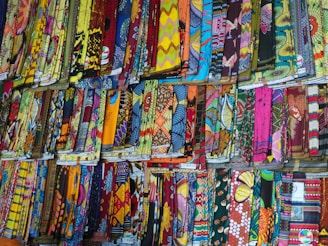 A display of various colorful, patterned fabrics arranged neatly in rows. The fabrics showcase an array of intricate designs, vibrant colors, and textures, creating a visually stimulating scene.