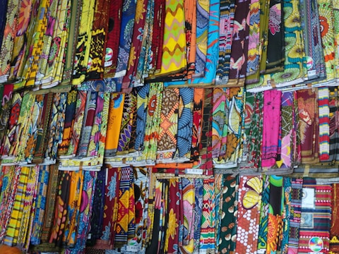 A display of various colorful, patterned fabrics arranged neatly in rows. The fabrics showcase an array of intricate designs, vibrant colors, and textures, creating a visually stimulating scene.