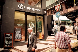A street scene featuring the entrance of a coffee shop named 'Cupping Room' with a prominent logo above the door and glass windows displaying the interior. A sandwich board on the sidewalk lists menu items. Two men are seen walking, one in a suit and another in casual clothing. A delivery truck is parked on the street, and there are several street signs, including a 'give way' sign.
