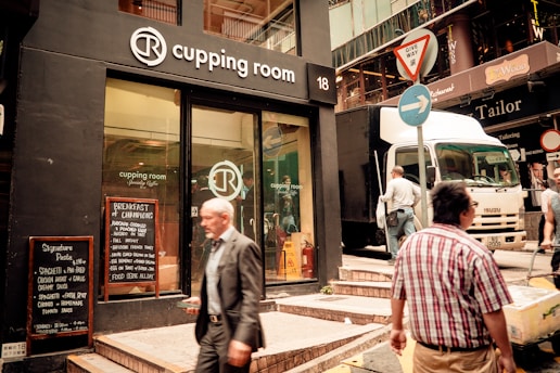 A street scene featuring the entrance of a coffee shop named 'Cupping Room' with a prominent logo above the door and glass windows displaying the interior. A sandwich board on the sidewalk lists menu items. Two men are seen walking, one in a suit and another in casual clothing. A delivery truck is parked on the street, and there are several street signs, including a 'give way' sign.