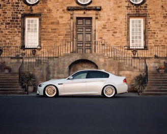A sleek white sedan is parked in front of an elegant brick building with large windows and a central door. The building's exterior features wrought iron railings and symmetrical staircases adorned with potted plants.