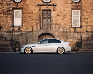 A sleek white sedan is parked in front of an elegant brick building with large windows and a central door. The building's exterior features wrought iron railings and symmetrical staircases adorned with potted plants.