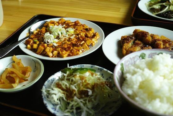 A variety of Asian dishes are presented on a wooden surface. The meal includes a plate of spicy tofu garnished with chopped green onions, crispy fried pieces, a bowl of plain white rice, a small plate of pickled vegetables, and a dish of shredded cabbage with sauce.