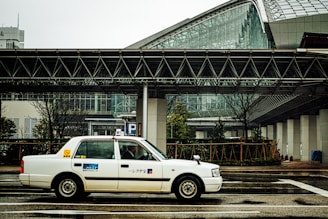 A white taxi is driving on a wet city street in front of a modern building with large glass windows and steel framework. The structure features geometric architectural elements, and there is a covered pedestrian walkway. Trees and some greenery are visible along the edge of the road.