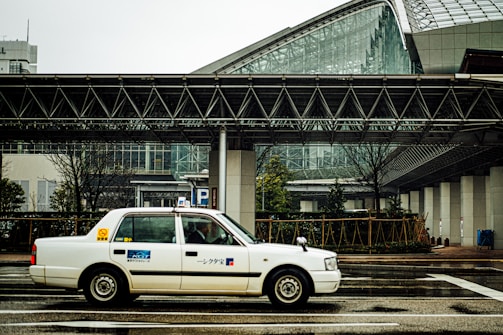 A white taxi is driving on a wet city street in front of a modern building with large glass windows and steel framework. The structure features geometric architectural elements, and there is a covered pedestrian walkway. Trees and some greenery are visible along the edge of the road.