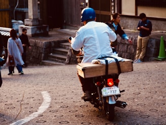 A person wearing a blue helmet and white jacket rides a motorbike with a wooden box attached to the back. The scene takes place on a street with cobblestone patterns, with several other people walking or standing nearby. Some are conversing while one individual appears to be using a mobile phone. The background features stone walls and steps.