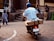 A person wearing a blue helmet and white jacket rides a motorbike with a wooden box attached to the back. The scene takes place on a street with cobblestone patterns, with several other people walking or standing nearby. Some are conversing while one individual appears to be using a mobile phone. The background features stone walls and steps.