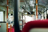 The interior of a bus with empty red seats and various handrails. The view through the front window is slightly blurred, and the bus driver is visible in the mirror at the front. The bus has a clean and modern design with informational signs and advertisements on the walls.