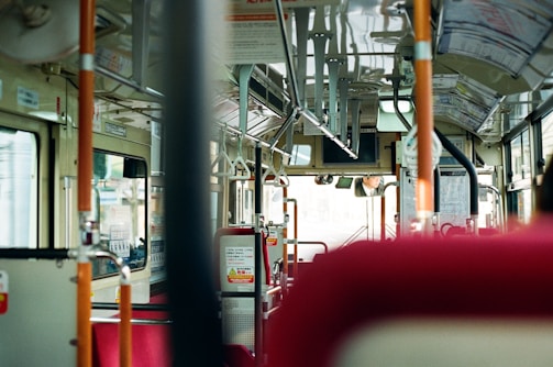 The interior of a bus with empty red seats and various handrails. The view through the front window is slightly blurred, and the bus driver is visible in the mirror at the front. The bus has a clean and modern design with informational signs and advertisements on the walls.