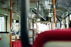 The interior of a bus with empty red seats and various handrails. The view through the front window is slightly blurred, and the bus driver is visible in the mirror at the front. The bus has a clean and modern design with informational signs and advertisements on the walls.