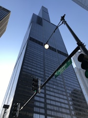 A tall, modern skyscraper with a glass facade extends upwards into the clear blue sky. The building is surrounded by other high-rise structures, and a street sign indicating Adams Street is visible in the foreground alongside traffic lights.
