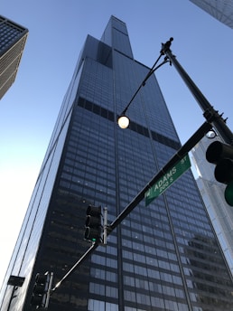 A tall, modern skyscraper with a glass facade extends upwards into the clear blue sky. The building is surrounded by other high-rise structures, and a street sign indicating Adams Street is visible in the foreground alongside traffic lights.