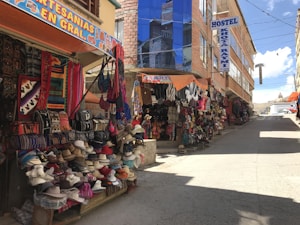 A street market with various colorful textiles and hats displayed outside stalls. The area is lined with buildings, one of which has a sign indicating a hostel. The scene is sunny with clear skies.