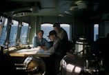 Three men are gathered around a table inside a ship's cabin, studying navigational charts. The room is dimly lit with natural light streaming through the windows, revealing a view of the ocean outside. Various maritime instruments and controls are visible, contributing to the nautical setting.