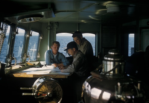 Three men are gathered around a table inside a ship's cabin, studying navigational charts. The room is dimly lit with natural light streaming through the windows, revealing a view of the ocean outside. Various maritime instruments and controls are visible, contributing to the nautical setting.