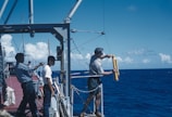Three individuals are on the deck of a research vessel in the ocean. One person is handling scientific equipment over the side of the ship, possibly preparing to deploy it into the water. The ocean is a deep blue, with a clear blue sky dotted with white clouds.