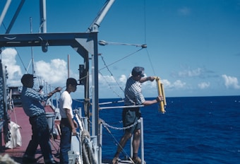 Three individuals are on the deck of a research vessel in the ocean. One person is handling scientific equipment over the side of the ship, possibly preparing to deploy it into the water. The ocean is a deep blue, with a clear blue sky dotted with white clouds.