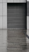 A modern, minimalist architectural facade featuring a series of horizontal metal slats within a concrete wall. The ground in front appears wet, reflecting the dull light and suggesting recent rain.