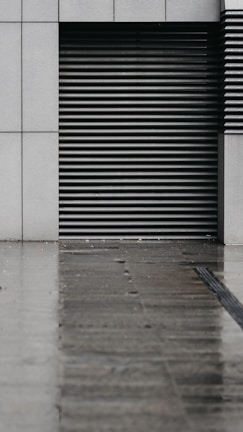 A modern, minimalist architectural facade featuring a series of horizontal metal slats within a concrete wall. The ground in front appears wet, reflecting the dull light and suggesting recent rain.