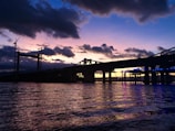Bridge under construction over a river with cranes and scaffolding at sunset.