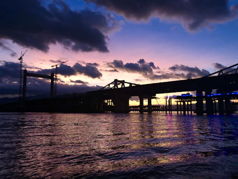 A panoramic view of a modern bridge construction site at sunset, showcasing cranes and workers.