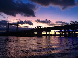 A partially constructed bridge extends over a body of water during a picturesque sunset. Two construction cranes are visible, silhouetted against the colorful sky. The clouds are dark, creating a contrast with the warm, glowing hues of orange, pink, and purple in the sky. The water below reflects these colors, adding to the serene yet industrial atmosphere.