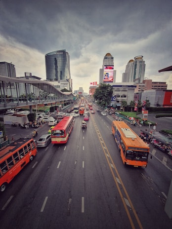 A busy urban street with multiple lanes of traffic, including several buses and cars, under a cloudy sky. Skyscrapers and a pedestrian overpass are visible in the background, and colorful billboards are displayed on the sides of buildings.