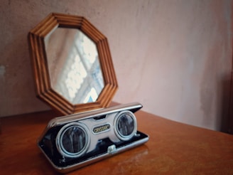 A wooden table with an octagonal mirror leaning against the wall and a vintage-style silver binocular case with the 'Crystar' brand name displayed on it, resting on the table.