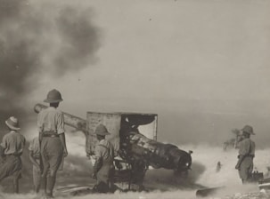 Several soldiers in uniforms and pith helmets operate and stand around a large piece of artillery. Smoke and dust fill the air, suggesting recent firing. The environment is sparse, and visibility is somewhat obscured by the smoke.