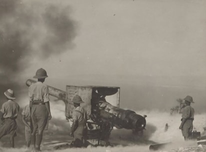 Several soldiers in uniforms and pith helmets operate and stand around a large piece of artillery. Smoke and dust fill the air, suggesting recent firing. The environment is sparse, and visibility is somewhat obscured by the smoke.
