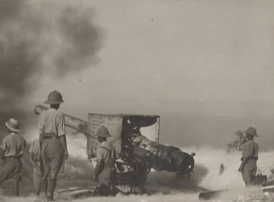 Several soldiers in uniforms and pith helmets operate and stand around a large piece of artillery. Smoke and dust fill the air, suggesting recent firing. The environment is sparse, and visibility is somewhat obscured by the smoke.
