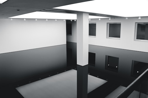 A minimalist interior space with a large open area resembling a gallery or atrium. The setting features a reflective black floor which gives an illusion of depth, and white walls adorned with evenly spaced windows. Overhead, a bright, evenly lit ceiling enhances the modern, clean lines of the architecture.