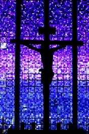 A silhouette of a crucifix is set against a backdrop of a stained glass window featuring a mosaic of deep blues and purples.